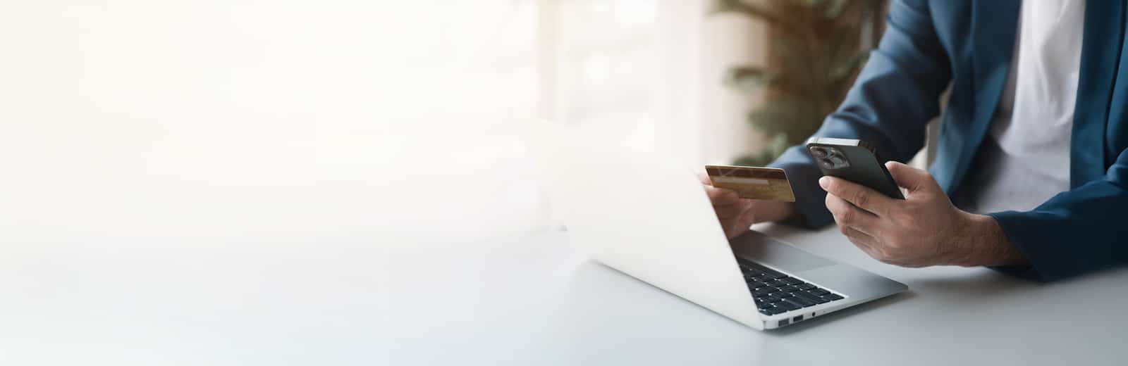 A person in a suit holds a smartphone and a credit card, seated at a bright desk with a laptop.