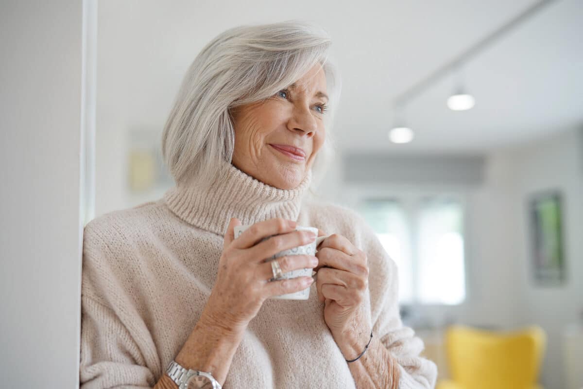 Elderly woman with gray hair in a cozy sweater, smiling softly while holding a cup in a bright living room. The atmosphere is warm and content.