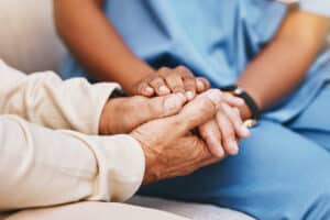 Two pairs of hands gently hold each other, conveying support and care. One person wears medical scrubs, suggesting a comforting healthcare setting.