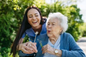 A joyful elderly woman blows a dandelion with the help of a smiling young caregiver outdoors.