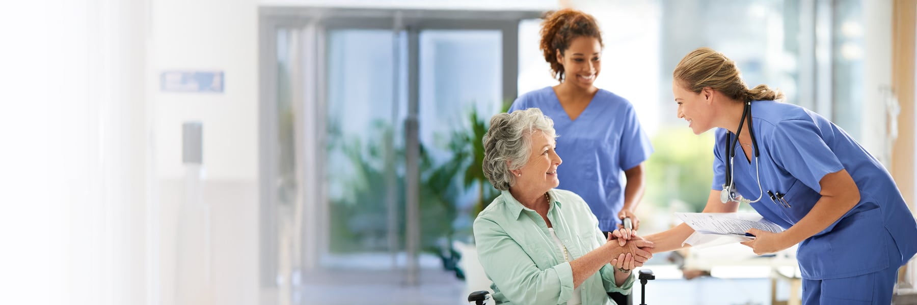 Nurses in blue scrubs warmly assist a smiling elderly woman in a wheelchair.