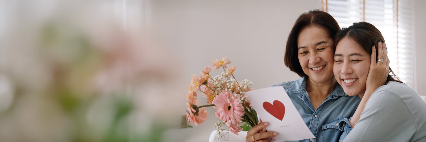 A woman and a young adult, both smiling warmly, embrace while holding a bouquet of pink flowers and a card with a red heart, conveying love and affection.