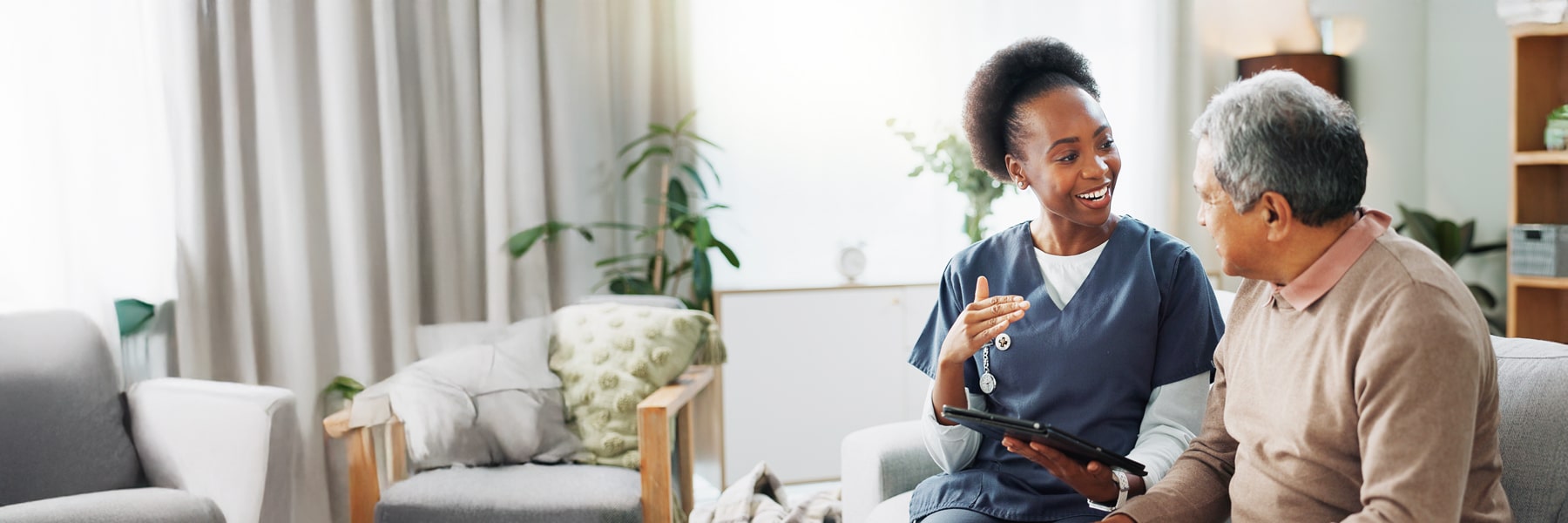 A nurse in blue scrubs is smiling and talking to an elderly man on a sofa in a cozy living room, holding a tablet.