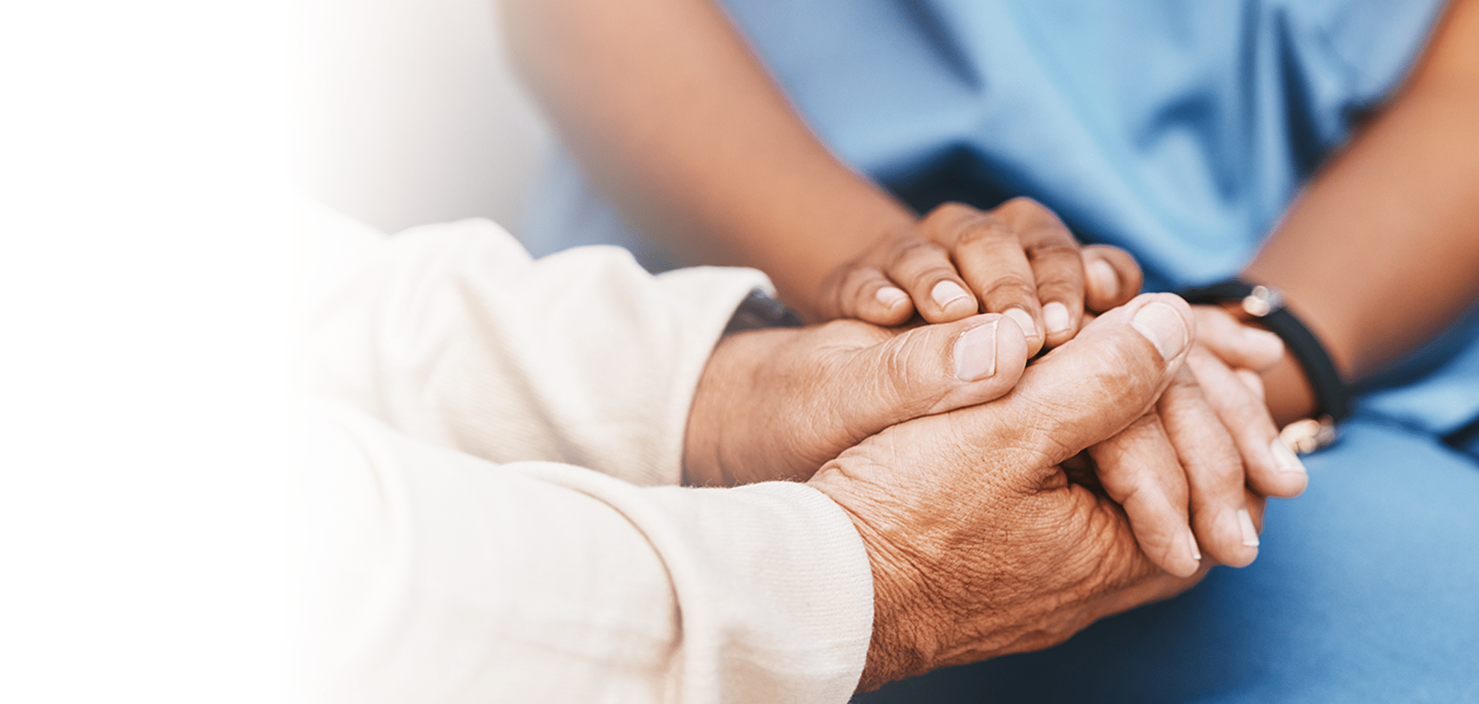 Two pairs of hands gently hold each other, conveying support and care. One person wears medical scrubs, suggesting a comforting healthcare setting.