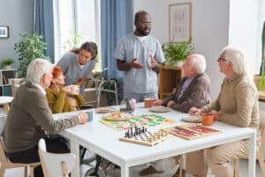 A group of elderly people sit around a table with board games, chatting happily with a caregiver in a bright, cozy room filled with plants and soft light.