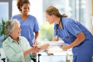 Nurses in blue scrubs warmly assist a smiling elderly woman in a wheelchair.