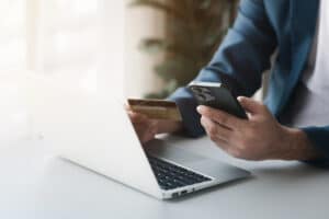 A person in a suit holds a smartphone and a credit card, seated at a bright desk with a laptop.