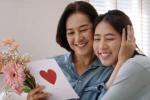 A woman and a young adult, both smiling warmly, embrace while holding a bouquet of pink flowers and a card with a red heart, conveying love and affection.