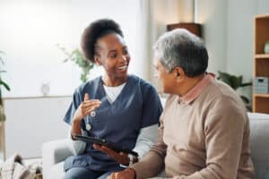 A nurse in blue scrubs is smiling and talking to an elderly man on a sofa in a cozy living room, holding a tablet.