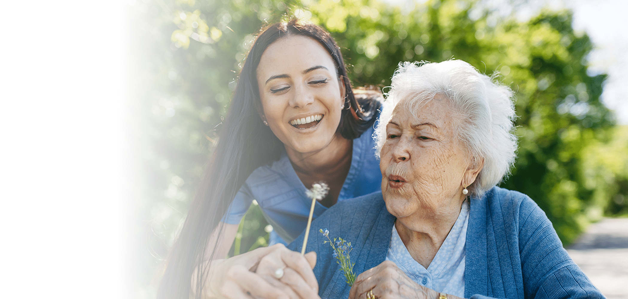 A joyful elderly woman blows a dandelion with the help of a smiling young caregiver outdoors.