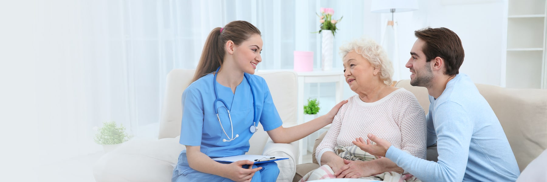 A nurse in blue scrubs warmly helps an elderly patient while a younger man is speaking to the nurse