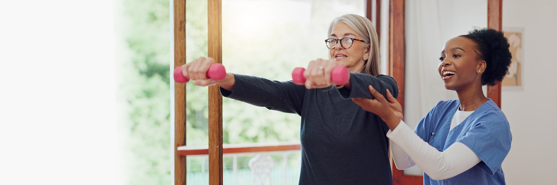 A woman is holding weights while a woman in nurse scrubs helps support her arms. They are in a bright gym, suggesting a supportive, focused rehabilitation session.