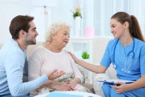 A nurse in blue scrubs warmly helps an elderly patient while a younger man is speaking to the nurse