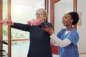 A woman is holding weights while a woman in nurse scrubs helps support her arms. They are in a bright gym, suggesting a supportive, focused rehabilitation session.